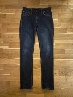 Mörkblå slim fit jeans från Nudie - Snygga mörkblå slim fit jeans från Nudie med klassiska fem fickor och kontrastsömmar. Jeansen har en smal passform och subtila slitningar framtill. Bakfickorna har Nudies ikoniska broderi. Perfekta för en stilren och avslappnad look.