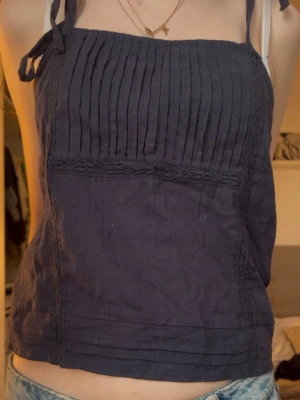 Mörkblå linnetopp med knytband - Snygg mörkblå linnetopp med knytbara axelband och ribbade detaljer framtill. Toppen är ärmlös och har en rak, något croppad passform. Perfekt för varma dagar och enkel att styla med jeans eller kjol. Går att justera armarna eftersom att man knyter. 
