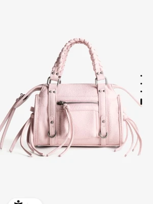 Ljusrosa bowlingväska med flätade handtag - Snygg ljusrosa handväska i läder med flätade handtag och silverfärgade metalldetaljer. Väskan har en framficka med dragkedja och dekorativa remmar på sidorna. Perfekt accessoar för att lyfta din outfit. Är använd ett fåtal gånger. Finns även med axelband. Originalpris; 329kr