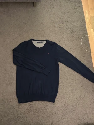 Mörkblå V-ringad tröja Tommy Hilfiger - Mörkblå V-ringad tröja från Tommy Hilfiger med diskret logga på bröstet. Tröjan har långa ärmar, ribbade muddar och är tillverkad i ett mjukt, tunt material som passar perfekt till lager-på-lager-stil.