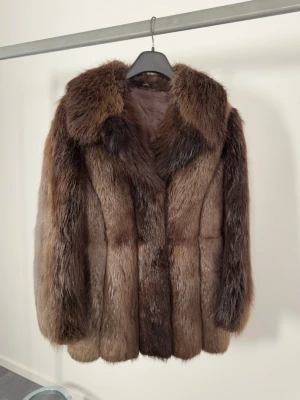 Äkta pälsjacka - Brun vintage pälsjacka i äkta päls. Rak modell med markerad krage och genomgående knäppning framtill (hooks/hyskor). Helfodrad insida. Längre modell som går ned över höften. Ärmar i full längd med generös vidd.  Utifrån utseende och hårstruktur bedöms den troligen vara räv, alternativt mårdhund. Exakt djurart kan inte garanteras utan märkning eller expertutlåtande.  Storlek: Uppskattas passa ca S–L beroende på önskad passform (oversized på mindre storlek). Axelvidd: ca. 50cm. Längd: ca. 75cm