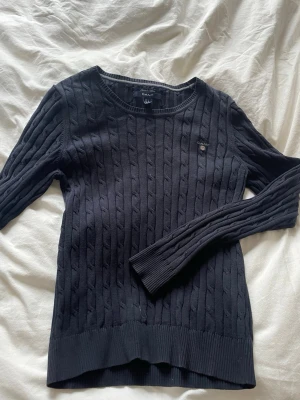 Mörkblå kabelstickad tröja GANT - Kabelstickad tröja från GANT i mörkblått med rund halsringning och diskret broderad logga på bröstet. Tröjan har långa ärmar och ribbade muddar vid ärmslut och nederkant. Perfekt för en klassisk och stilren look.