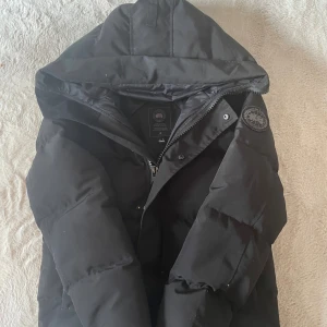 Svart dunjacka från Canada Goose - Snygg svart dunjacka från Canada Goose med huva och klassisk patch på ärmen. Jackan har dragkedja och tryckknappar framtill, perfekt för kalla vinterdagar. Stilren och tidlös design som håller dig varm.  Vill lägga till att den tyvärr tappar fjädrar på vissa stället. Går ej att fixa det utan den är så drf säljer jag billigt