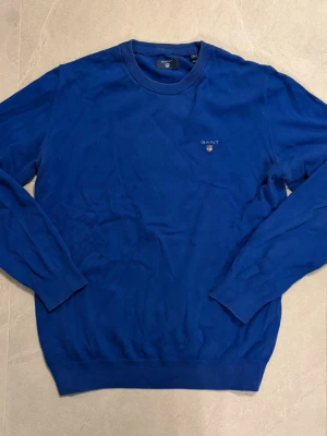 Blå stickad tröja från Gant - Klassisk blå stickad tröja från Gant med rund halsringning och diskret logga broderad på bröstet. Tröjan har långa ärmar och ribbade muddar vid ärmslut och nederkant. Perfekt för en stilren och avslappnad look.