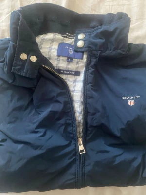 Marinblå vindjacka från GANT - Snygg marinblå vindjacka från GANT med dragkedja framtill, två fickor med tryckknappar och broderad logga på bröstet. Jackan har ståkrage med knappar och rutigt innerfoder. Perfekt för blåsiga dagar och enkel att matcha med olika outfits.