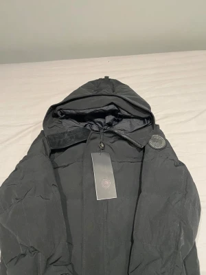 Svart pufferjacka med huva - Säljer en svart pufferjacka från Peak Performance med stor huva och dragkedja framtill. Jackan har en diskret logotyp på ärmen och är tillverkad i ett slitstarkt, vadderat material som håller värmen under kalla dagar. Perfekt för dig som vill ha en stilren och funktionell vinterjacka.