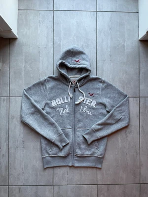 Grå Hollister hoodie med dragkedja - Snygg grå hoodie från Hollister med dragkedja framtill och broderad text 'Hollister Malibu' i vitt. Klassisk huva med dragsko och Hollister-logga i rött på bröstet. Mjuk bomullsblandning och ribbade muddar vid ärmar och nederkant.