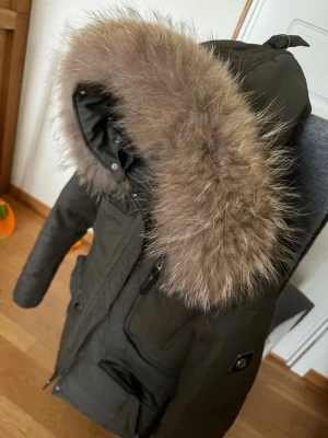 Olivgrön parkas från Hollies med pälskrage - Snygg olivgrön parkas från Hollies med stor fluffig pälskant på huvan. Jackan har dragkedja, flera fickor och en klassisk längre modell. Perfekt för kalla dagar och riktigt trendig look. Materialet är slitstarkt och fodrat för extra värme. Jackan är i storlek 38 men mer som en 36