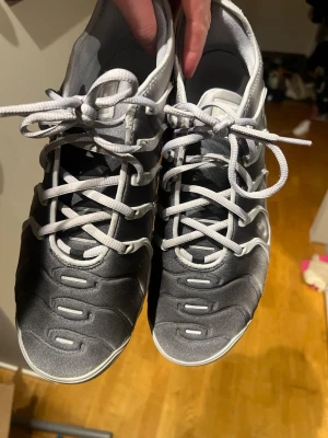 Nike Air Vapormax Plus - Äkta Nike Air Vapormax Plus sneakers i en snygg grå och svart färgskala med vita detaljer. Skorna har en unik plaststruktur på ovansidan, genomskinlig sula med synliga luftbubblor och grå skosnören. Perfekt för dig som vill ha en modern och sportig look.