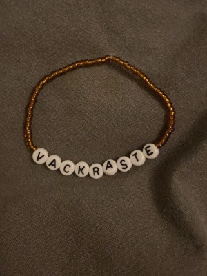 Pärlarmband med texten VACKRASTE - Ett handgjort armband med små guldfärgade pärlor och vita runda bokstavspärlor som bildar ordet 'VACKRASTE'. Perfekt accessoar för att ge en personlig touch till din stil.