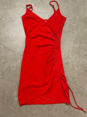 Röd omlottklänning från Divided - Snygg röd kortklänning från Divided (hm) med omlott-design och smala axelband. Klänningen har en draperad detalj med snörning på sidan som ger en cool och trendig look. Perfekt för dig som vill sticka ut med en färgstark outfit.           Obs fläck bild 3. Kan mötas upp i Stockholm.
