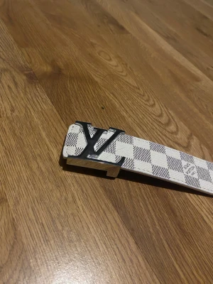 Rutigt bälte i vitt - Snyggt bälte med klassiskt rutigt mönster i vitt och beige. Bältet har en stor logga i metall som spänne och är tillverkat i skinn. Perfekt accessoar för att lyfta din outfit.