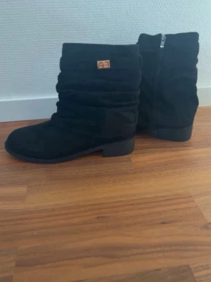 Svarta slouchy stövlar med dragkedja - Säljer ett par svarta stövlar i mockaimitation med slouchy design och låg klack. Stövlarna har dragkedja på sidan och en liten gulddetalj upptill. Perfekta till höstens outfits och ger en avslappnad men snygg look.