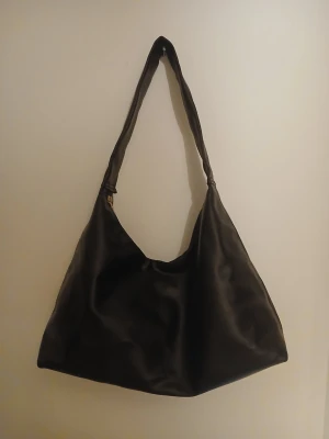 Svart axelväska i fejkläder - Stilren svart axelväska i mjukt fejkläder med enkel, slouchy design och bred axelrem. Väskan har guldfärgad metalldetalj vid remmen och rymlig insida. Perfekt för dig som gillar minimalistisk och trendig look.