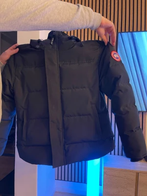 Svart pufferjacka från Canada Goose - Säljer en svart pufferjacka från Canada Goose med klassisk patch på ärmen. Jackan har huva, hög krage och breda kanaler för extra värme. Perfekt för kalla vinterdagar och riktigt snygg till streetwear-stilen.