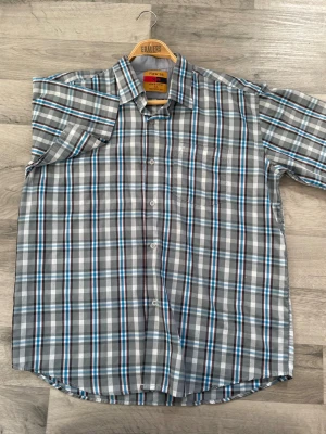 Rutig kortärmad skjorta från Raw-18 - Snygg kortärmad skjorta från Raw-18 med rutigt mönster i grått, vitt, blått och svart. Skjortan har klassisk krage, knäppning framtill och bröstficka. Perfekt för en avslappnad stil.