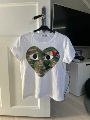 Comme des Garçons Play t-shirt - Vit t-shirt från Comme des Garçons Play med stort hjärta i camouflage-mönster och ögon på bröstet👌 Liten röd hjärt-detalj med ögon på vänster sida. Fint skick i storlek S.
