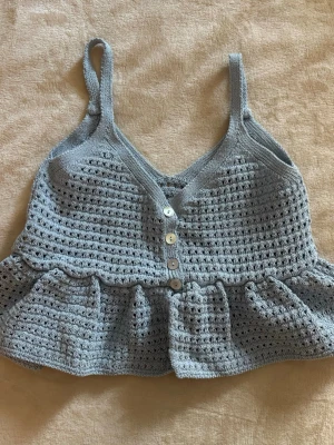 Ljusblå stickad topp med knappar - Supersöt ljusblå virkad/stickad topp med smala axelband och volang nertill. Toppen har v-ringning och dekorativa knappar framtill. I storlek S men passar Xs precis lika bra🤍