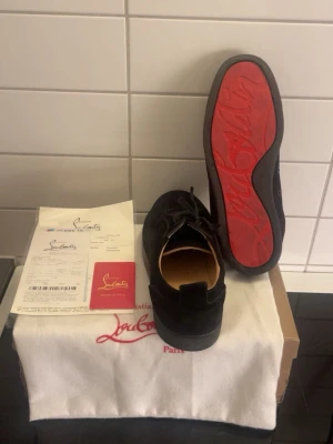 Svarta sneakers från Christian Louboutin - Snygga svarta sneakers från Christian Louboutin med den ikoniska röda sulan och diskret logga. Skorna har en klassisk låg siluett, snörning framtill och är tillverkade i mocka. Perfekta för dig som vill ha något exklusivt och stilrent.