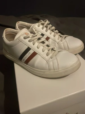 Vita Moncler sneakers  - Snygga vita sneakers från Moncler i läder med klassiska ränder i svart, rött och vitt på sidan. Skorna har lågt skaft, vita snören och diskret Moncler-logga. Perfekta för dig som gillar stilrena och exklusiva sneakers. Box med kommer 