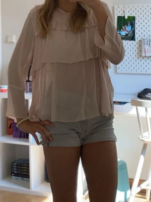 Ljusrosa volangblus med långa ärmar neo nor  - Söt ljusrosa blus med volangdetaljer och lös passform. Blusen har långa ärmar och är tillverkad i ett tunt, lätt material som ger en luftig känsla. Perfekt för en trendig och avslappnad look.