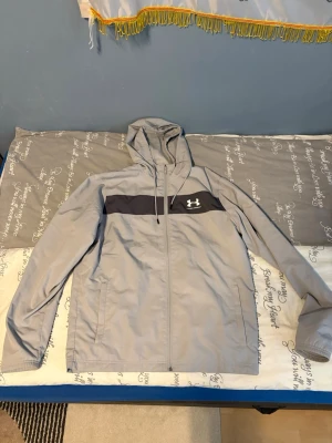Grå vindjacka från Under Armour - Snygg grå vindjacka från Under Armour med svart detalj över bröstet och logga. Jackan har huva med dragsko, hel dragkedja och två sidofickor. Tillverkad i lätt och ventilerande material med meshfoder för extra komfort.