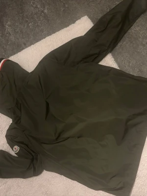 Moncler grön vindjacka med huva - Stilren grön vindjacka från Moncler med huva och dragkedja framtill. Jackan har en vit och röd rand på huvan samt Moncler-logga på bröstet. Perfekt för blåsiga dagar och enkel att matcha med olika outfits. Jackan är 100% ÄKTA säljer pga passar inte mig. Skriv privat för mer bilder