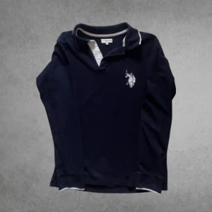 Polo piké - Snygg marinblå långärmad pikétröja från U.S. Polo Assn med vit broderad logga på bröstet. Tröjan har klassisk krage, vita detaljer vid knäppningen och muddar vid ärmslut. Perfekt för dig som gillar sportig och stilren look.