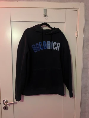 Mökrblå hoodie från Hoodrich  - Mörkblå hoodie från Hoodrich med stor blå och vit logotyp på bröstet. Klassisk huva med dragsko och en stor magficka framtill. Perfekt för dig som gillar streetwear och vill ha en snygg och bekväm hoodie med coolt tryck. Skicket på den är bra, hör av er om ni har frågor.