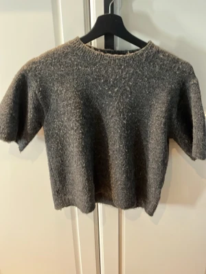 Mörkgrå stickad tröja Lindex - Mysig mörkgrå stickad tröja från Lindex med korta ärmar och rund hals. Tröjan har en enkel, rak passform och är tillverkad i ett mjukt, fluffigt material som känns skönt mot huden. Perfekt för lager på lager och en avslappnad look.