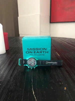 Swatch x Omega Mission on Earth klocka - Cool Swatch x Omega Speedmaster Mission on Earth Polar Lights klocka i turkos med svart textilband och kontrastsömmar. Klockan har kronograf och detaljerad urtavla, samt en bild av jorden på baksidan. Perfekt för dig som gillar unika accessoarer. Köpt för 6000 när klockan släpptes sänkt i pris nu en mycket fin Limiterad klocka som Komemr ökas mycket i pris först till kvarn 😁