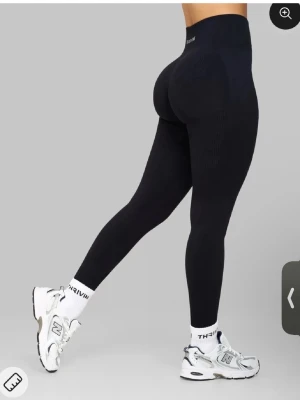 Svarta seamless leggings från Thrive - Snygga svarta seamless leggings från Thrivin med hög midja och tight passform. Materialet är stretchigt och formar sig efter kroppen, perfekt för träning eller chill. Diskret logga i midjan och ribbade detaljer längs benen för extra stil.