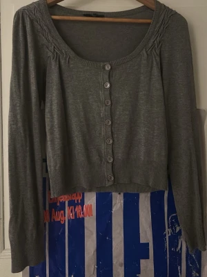 Grå cropped kofta med knappar - Snygg grå kofta med långa ärmar och rundad urringning. Koftan har dekorativa veck vid axlarna och stängs med flera knappar framtill. Perfekt att styla över ett linne. Vet tyvärr inte var den är ifrån då lappen är bortklippt