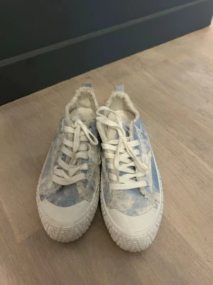 Blå tie-dye sneakers med fransade kanter - Sneakers i blått och vitt tie-dye-mönster med fransade kanter och vita snören. Skorna har en chunky vit sula med mönster och metallfärgade öljetter. Tillverkade i canvasmaterial för en avslappnad vibe.