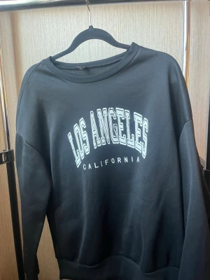 Svart sweatshirt Los Angeles-tryck - Svart sweatshirt med vitt tryck där det står 'Los Angeles California' på bröstet. Tröjan har rund halsringning och långa ärmar. 