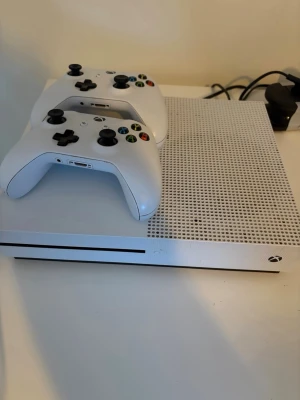 Xbox One S med två vita handkontroller - Säljer en Xbox One S i vit färg med två matchande vita handkontroller. Konsolen har en stilren, rektangulär design med perforerad ovansida. Medföljer flera populära spel som FIFA, Call of Duty, Battlefield och GTA V. Perfekt för gaming och multiplayer.