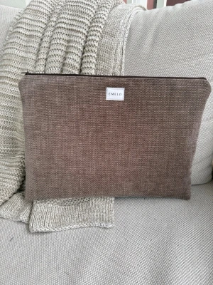 Brun datorväska i tyg från Emelo - Stilren datorväska från Emelo i brunt möbeltyg och dragkedja upptill. Väskan har en enkel, rektangulär form och en diskret logotyp framtill. Perfekt för att bära din laptop på ett snyggt och praktiskt sätt. Vadderat. Passar laptop med maxmått: 33x24cm