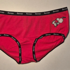 Röda Snoopy trosor med tryck storlek M - Säljer ett par röda trosor med svart resår och Snoopy-tryck både fram och bak. Resåren har texten 'Snoopy' runtom och trosorna har en klassisk brief-modell. Materialet känns mjukt och stretchigt, troligen bomullsblandning. Perfekta för dig som gillar söta och lekfulla detaljer. Storlek M, aldrig använda. Kommer från konkurslager. Bundlerabatt på 20% vid flerköp.