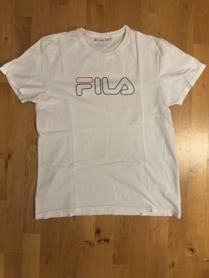 Vit FILA t-shirt med logga - Klassisk vit t-shirt från FILA med stor logga i svart och rött framtill. T-shirten har rund halsringning och korta ärmar. Tillverkad i mjuk bomull för en skön känsla och enkel stil.