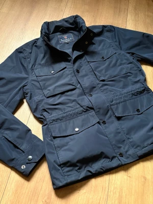 Bondelid field jacket navy - Snygg mörkblå vindjacka från Bondelid med hög krage och flera praktiska fickor framtill. Jackan har tryckknappar, dragkedja och justerbara ärmslut. Tillverkad i ett lätt, vattenavvisande material som passar perfekt för blåsiga dagar.
