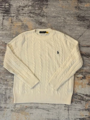 Vit kabelstickad tröja Polo Ralph Lauren - Kabelstickad vit tröja från Polo Ralph Lauren med rund halsringning och broderad marinblå logga på bröstet. Tröjan har ribbade muddar och nederkant, klassisk passform och är tillverkad i mjukt material.