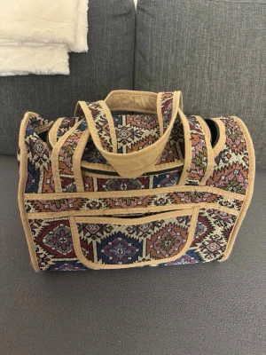 Mönstrad weekendbag i gobelängtyg - Säljer en unik resväska i gobelängtyg med orientaliskt mönster i färgerna beige, blå, lila och vinröd. Väskan har mjuka handtag och beigea kanter. Perfekt för kortare resor eller som snygg accessoar på tågresan.