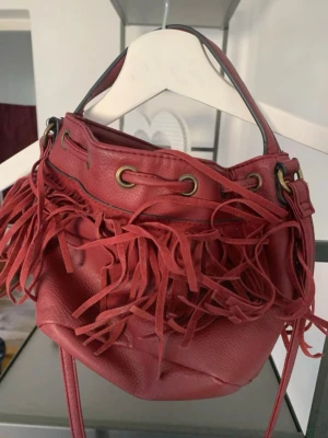 Vinröd handväska med fransar - Cool vinröd handväska i mjukt skinnimitation med långa fransar och dragskostängning. Väskan har både handtag och axelrem samt detaljer i mässingsfärgad metall. Perfekt för dig som vill sticka ut med en unik accessoar.