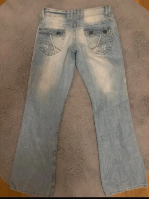 Ljusblå bootcut jeans House of denimn - Säljer ett par ljusblå bootcut jeans från House of denimen med slitna detaljer och dekorativa bakfickor med knappar. Jeansen har låg midja och klassisk femficksdesign. Materialet är denim i bomull och passformen är avslappnad. Supersöta!