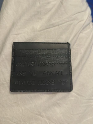 Svart korthållare från Armani Jeans - Snygg svart korthållare från Armani Jeans i slimmat format. Tillverkad i präglat läder med logotyp och ikoniska Armani-mönster över hela ytan. Flera fack för kort på båda sidor, perfekt för dig som vill ha stilren och praktisk accessoar.             (Priset är diskuterbart)