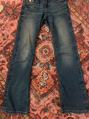 jeans från Gina  - Jeansen har framfickor och bakfickor med lock och knappdetaljer. Tillverkade i mjukt denimtyg som ger en skön passform och avslappnad look.