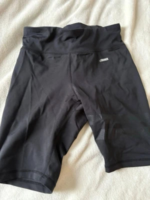 Svarta träningstights-shorts STRONGER - Säljer ett par svarta träningstights-shorts från STRONGER i storlek M. De har hög midja och diskreta meshdetaljer på benen för extra ventilation. Perfekta för gymmet eller löprundan och sitter riktigt skönt på kroppen.