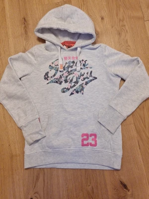 Ljusgrå hoodie från Superdry - Ljusgrå hoodie från Superdry med tryckt logga i blommigt mönster på bröstet och siffran 23 i rosa nedtill. Huva med vita dragsnören och magficka framtill. Perfekt för en avslappnad och trendig stil.
