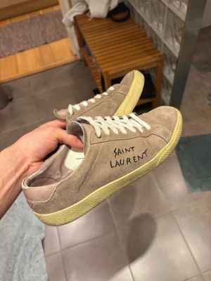 Saint Laurent beige mockasneakers - Snygga beige sneakers från Saint Laurent i mocka med vit sula och vita skosnören. Klassisk låg modell med handskriven logga på sidan. Perfekta för dig som gillar stilrena och exklusiva sneakers med en unik touch.