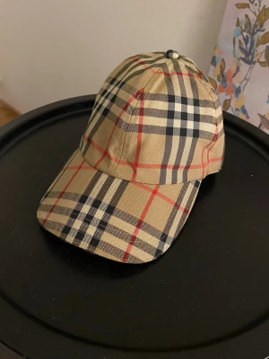 Burberry keps med klassiskt rutmönster - Snygg keps från Burberry i beige med det ikoniska rutmönstret i svart, vitt och rött. Kepsen är tillverkad i bomull och har böjd skärm. Perfekt accessoar för att lyfta din outfit med en lyxig touch.